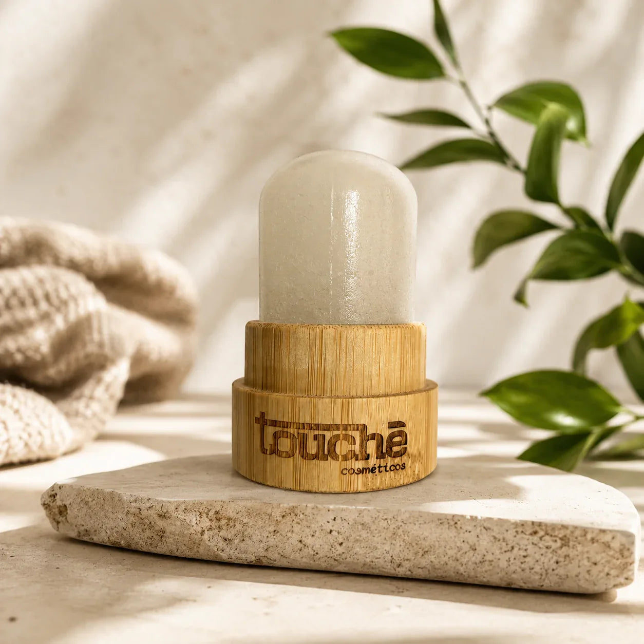 Desodorante natural em bastão com embalagem de bambu da Touché Cosméticos, sobre pedra com folhas verdes ao fundo